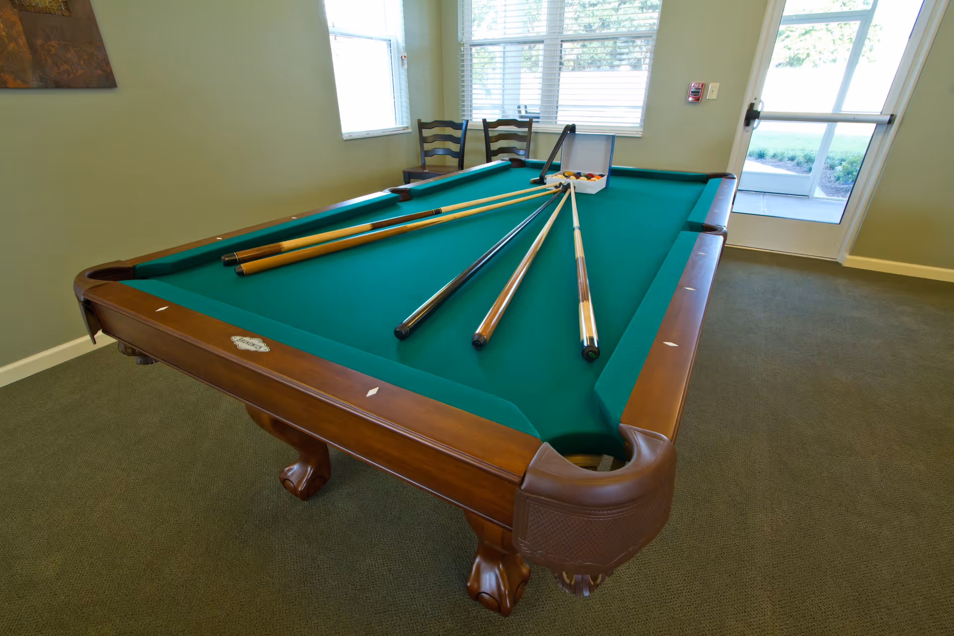 A billiards room with a green felt pool table, several pool cues resting on the table, and a rack of billiard balls. The room has green walls, carpeted floor, two wooden chairs near the window, and a glass door leading outside.
