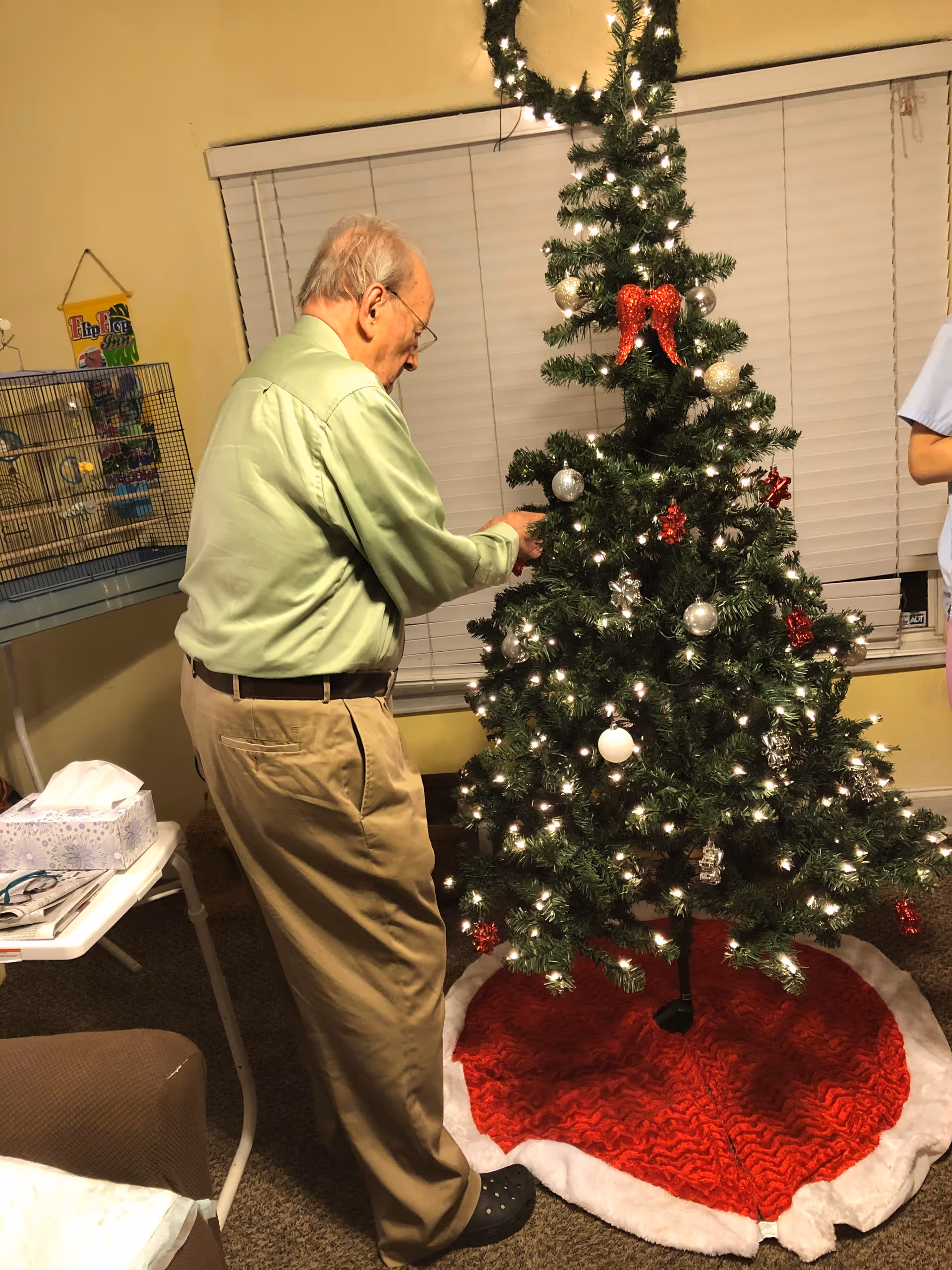 An elderly man wearing a light green shirt and beige pants decorates a Christmas tree with white and red ornaments in a cozy room with yellow walls and closed window blinds. A birdcage and a small table with tissues and reading glasses are visible nearby.