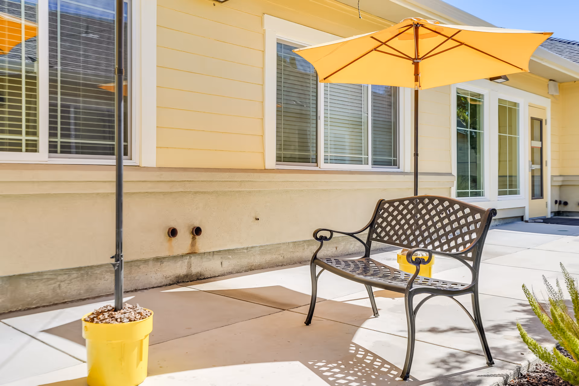 Outdoor patio area with a metal bench and a yellow umbrella providing shade. The patio is adjacent to a yellow building with multiple windows and a door. There are two yellow pots with umbrellas on the concrete floor.
