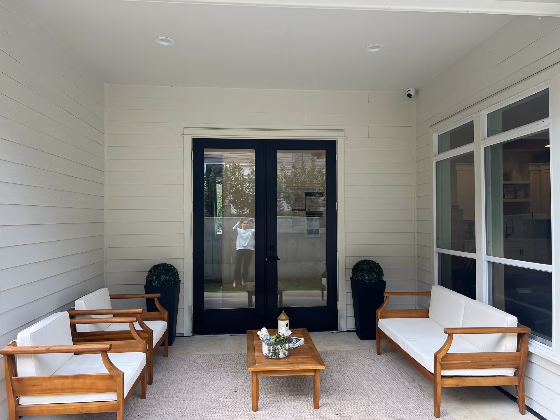 Covered porch with wooden-framed white-cushioned seating and a central coffee table facing black double glass doors.