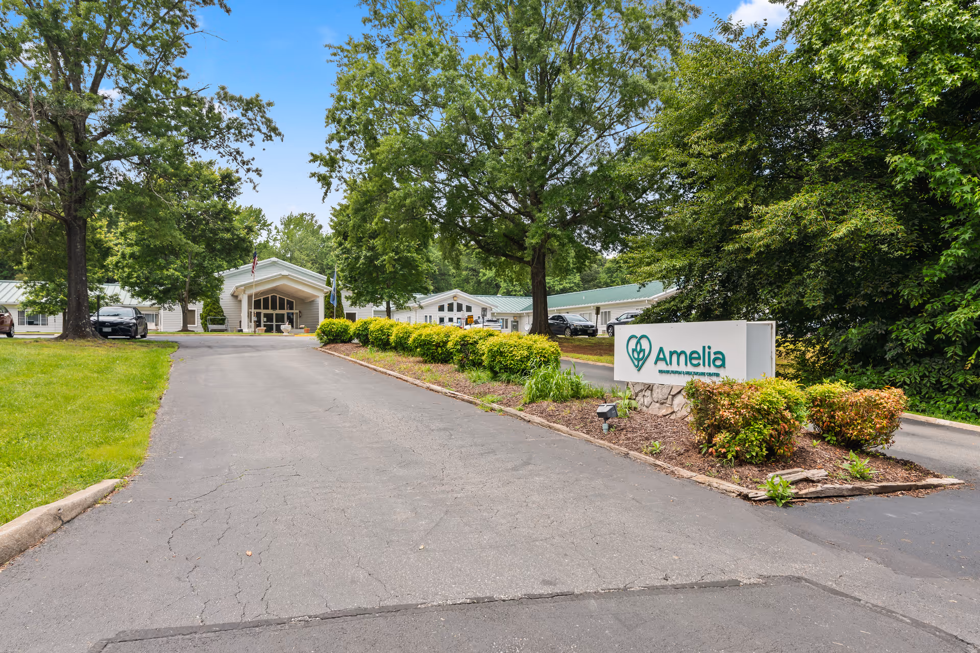 Entrance driveway leading to a single-story building surrounded by green trees and bushes. A sign on the right side reads 'Amelia Rehabilitation and Healthcare Center'. Several cars are parked near the building under a clear blue sky.