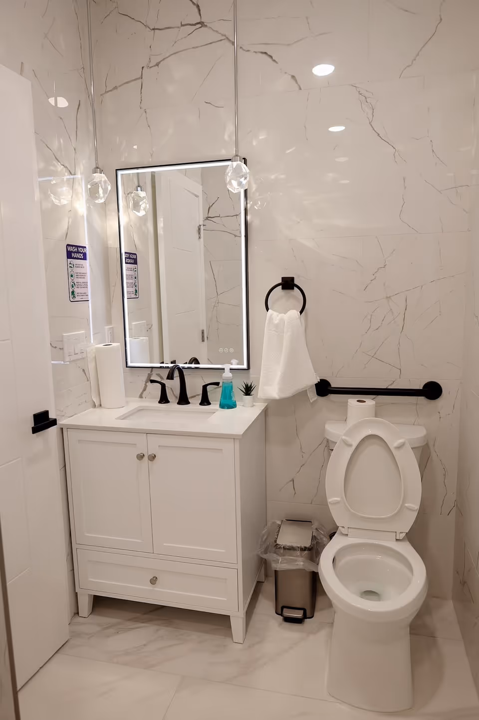 A clean, modern bathroom with white marble-patterned walls and floor. It features a white vanity with a sink, black faucet, a rectangular mirror with built-in lighting, two hanging pendant lights, a towel ring with a white towel, a toilet with the lid open and a roll of toilet paper on top, a small trash can with a plastic liner, and a soap dispenser on the vanity.