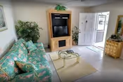 A bright living room with a floral patterned green sofa, a glass coffee table on a beige rug, a wooden entertainment center with a TV, potted plants, and a white front door with windows letting in natural light.