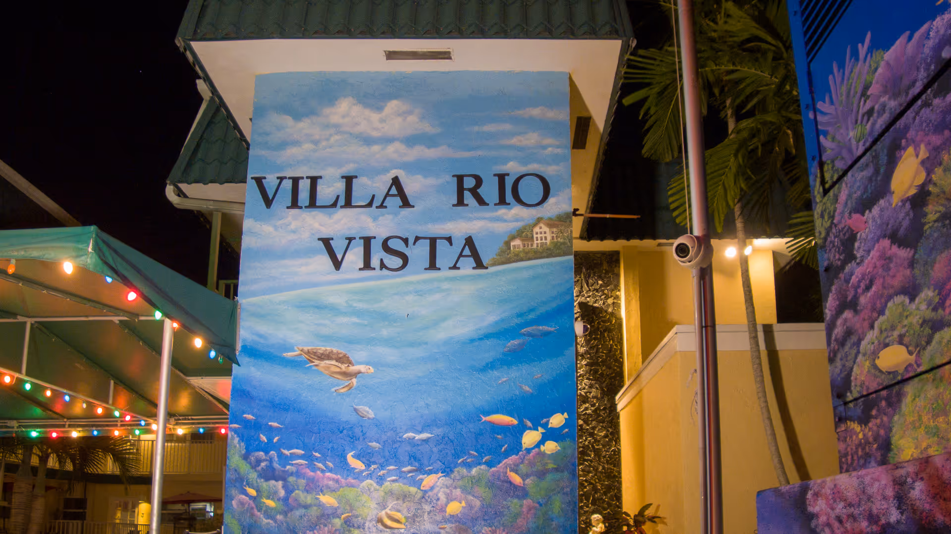 A painted mural on a building wall at night displaying the words 'VILLA RIO VISTA' with an underwater ocean scene featuring a sea turtle swimming among colorful fish and coral. The scene is illuminated by nearby lights and there are palm trees and a green roof visible in the background.