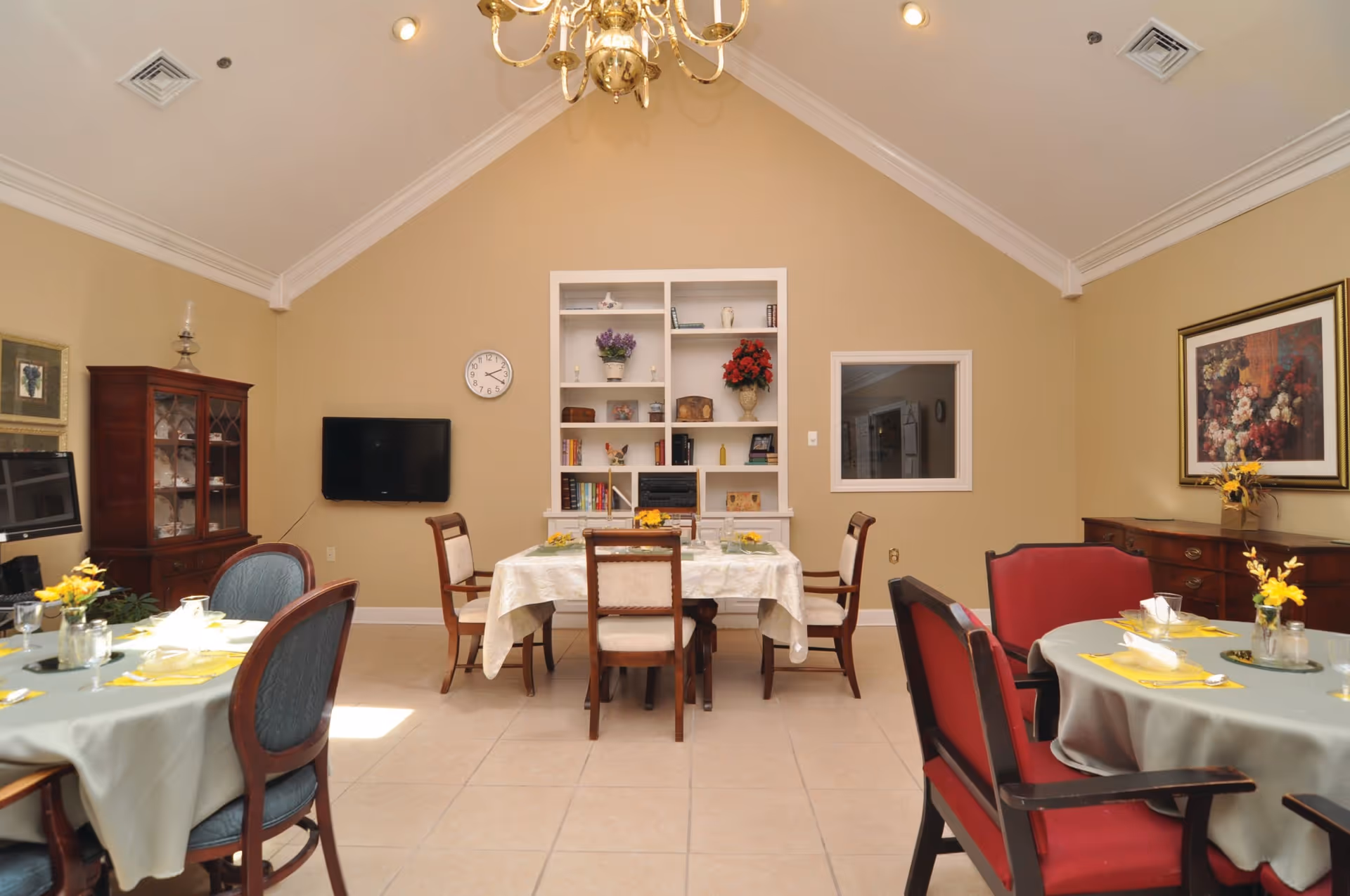 A dining room in Rose Wood Manor with several tables covered with tablecloths and set with plates, glasses, and yellow napkins. The room has beige walls, a high vaulted ceiling with a chandelier, a wall-mounted TV, a clock, a white built-in bookshelf with decorative items, and framed artwork on the walls. There are wooden chairs with upholstered seats around the tables.