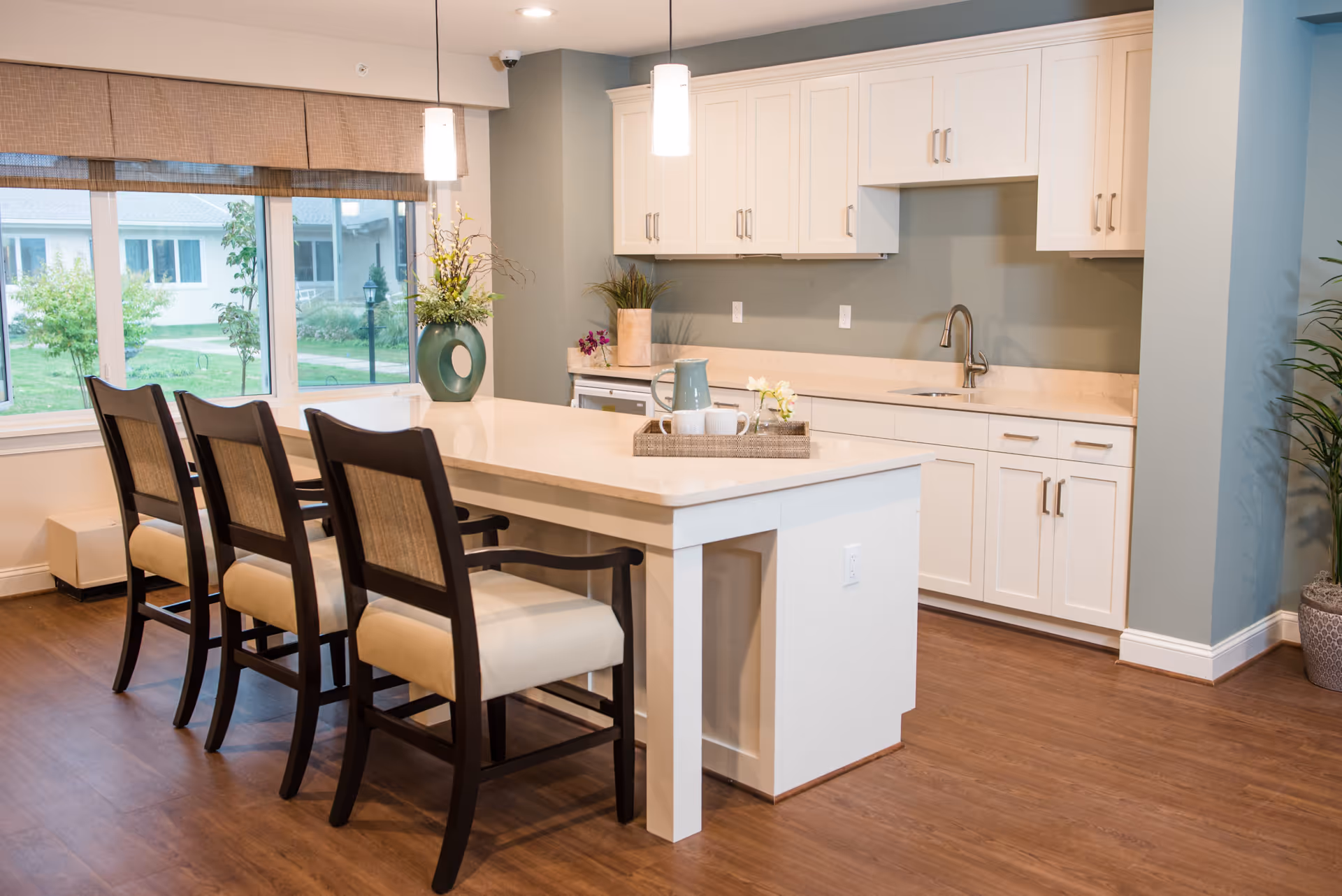 A modern kitchen area with a large white island countertop surrounded by three wooden chairs with beige cushions. The kitchen has white cabinets, a sink with a faucet, and two pendant lights hanging above the island. A large window with beige blinds shows a green outdoor area. Decorative plants and a tray with a pitcher and cups are placed on the countertop.