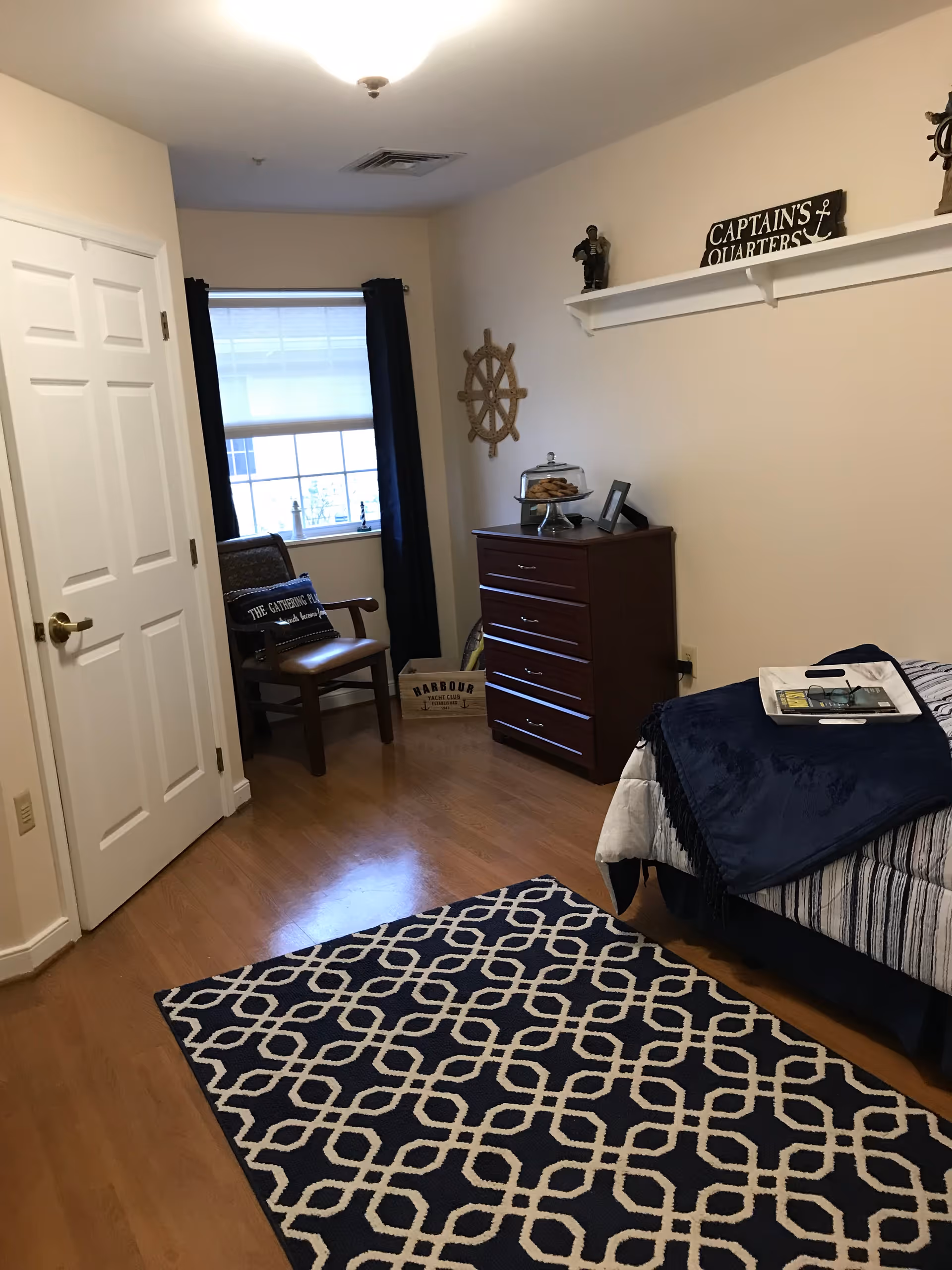 A cozy bedroom with a wooden floor, a patterned navy and white rug, a bed with a navy blanket and a tray on top, a wooden dresser with a glass dome containing cookies, a chair with a cushion near a window with dark curtains, and nautical-themed decorations including a ship's wheel and a sign that reads 'Captain's Quarters'.