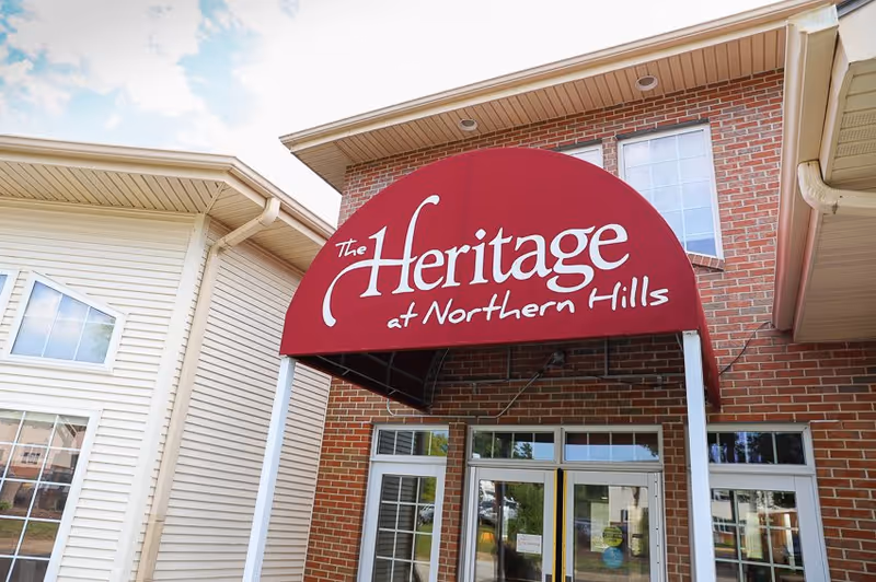 Entrance of The Heritage at Northern Hills facility showing a red awning with the facility name in white text above glass double doors, with brick and siding exterior walls.