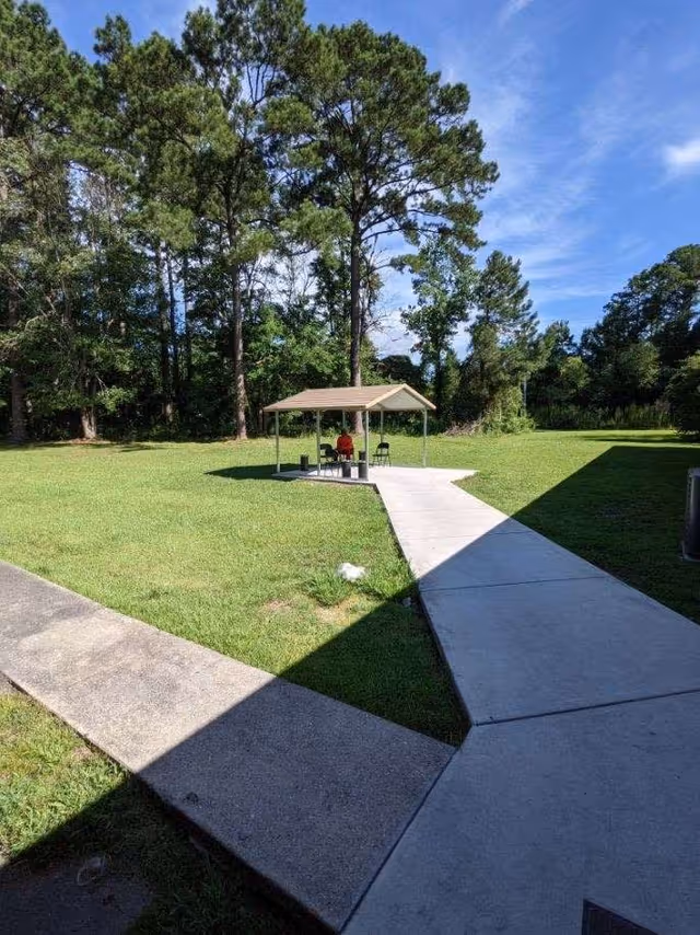 A small covered seating area with a bench and two chairs is situated on a concrete path in a grassy outdoor space surrounded by tall trees under a blue sky with some clouds.