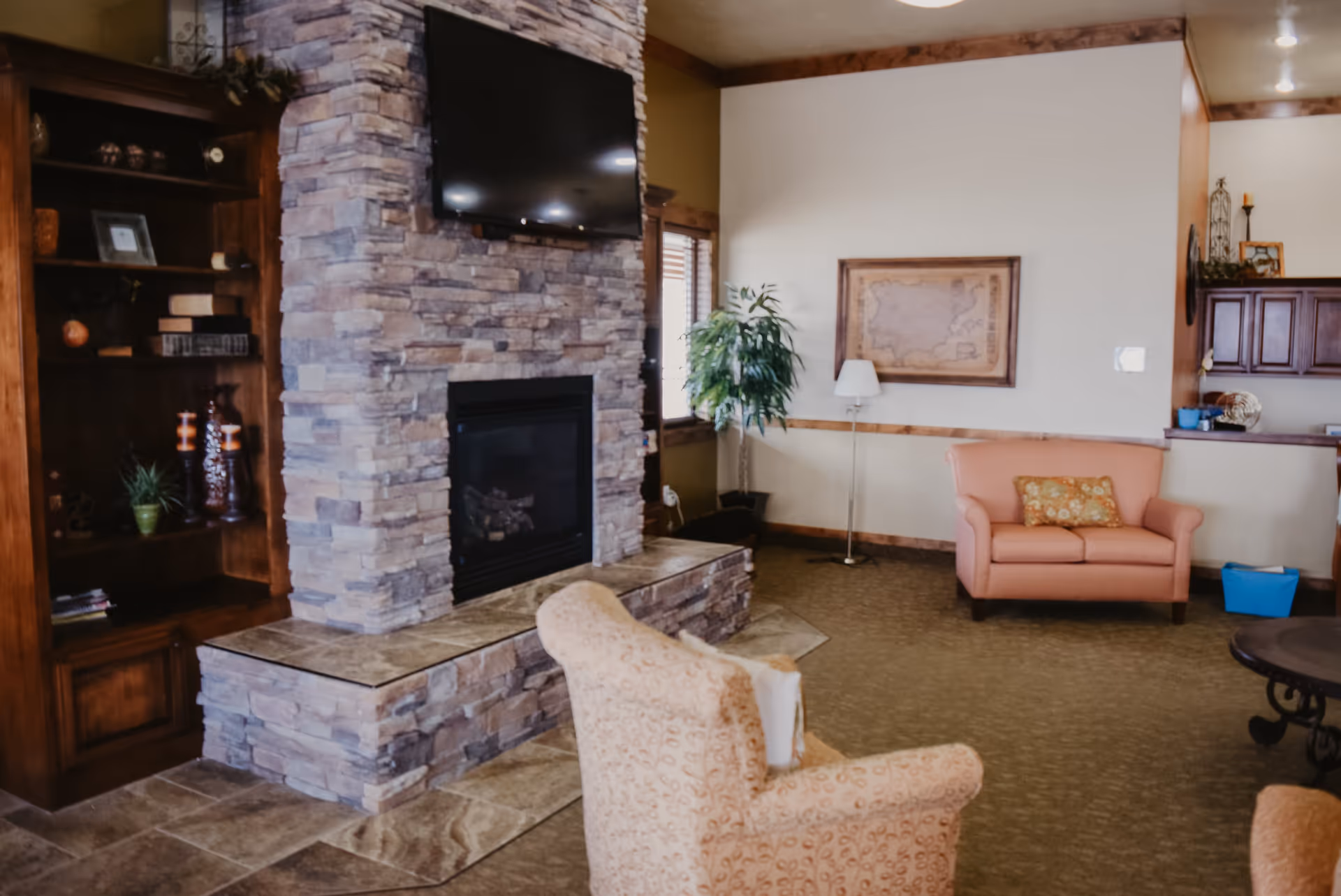 A cozy living room area with a stone fireplace and a mounted flat screen TV above it. There is a wooden bookshelf with decorative items and books on the left side of the fireplace. In the foreground, there is a patterned armchair, and in the background, a peach-colored loveseat with a decorative pillow. A floor lamp and a framed map hang on the wall behind the loveseat. The room has carpeted flooring and wooden trim accents.