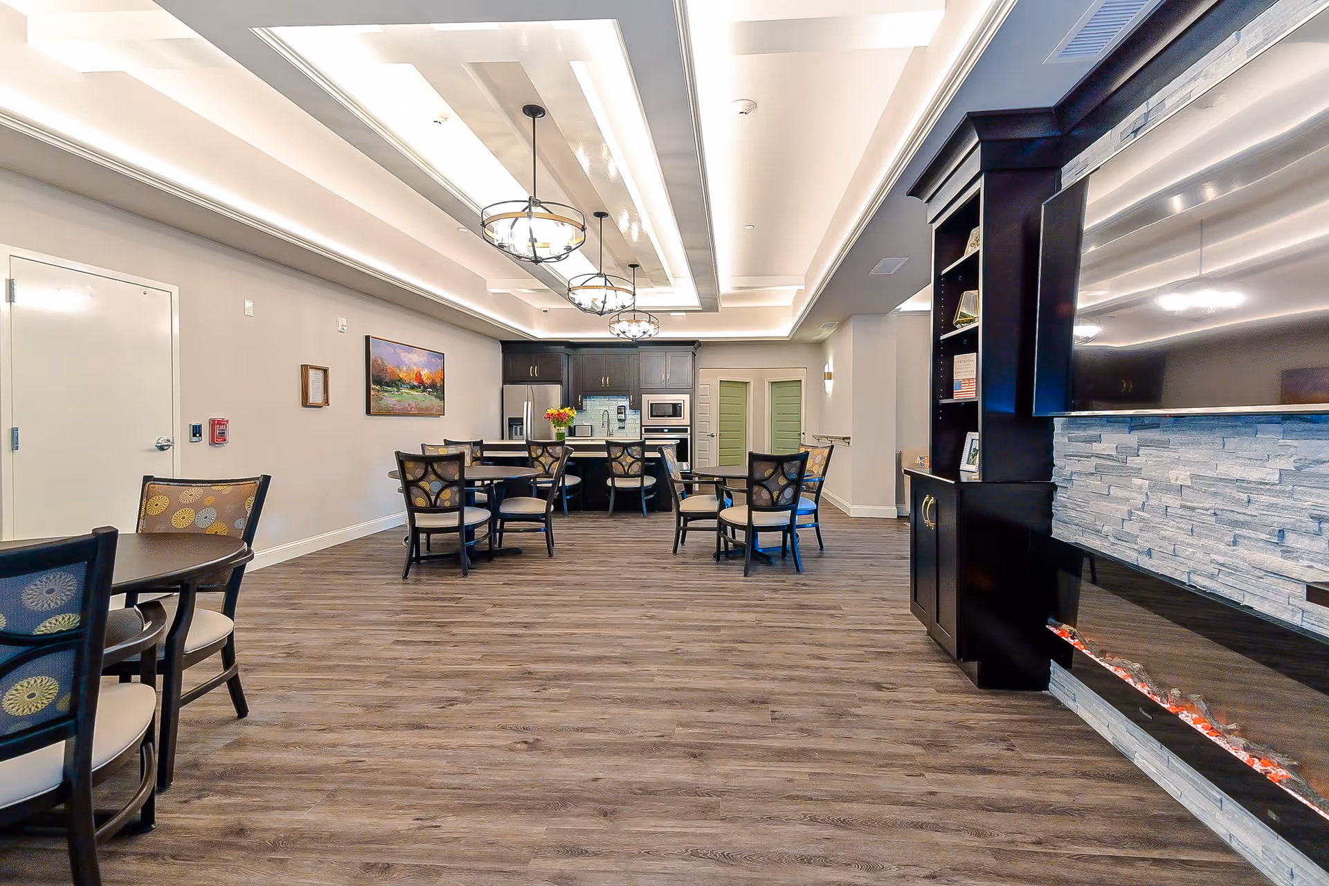Communal dining and lounge area with round tables and chairs, a kitchen at the back, and a modern fireplace on the right.