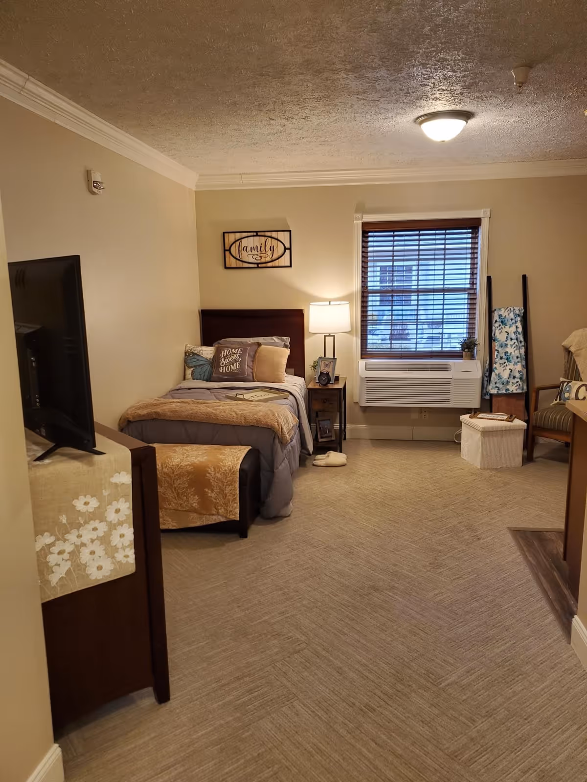 A cozy bedroom in a senior living facility with a single bed covered in gray and beige bedding, a small wooden nightstand with a lamp and picture frame, a window with blinds and an air conditioning unit below it, a TV on a stand with a floral cloth, and a chair with a blanket draped over it.