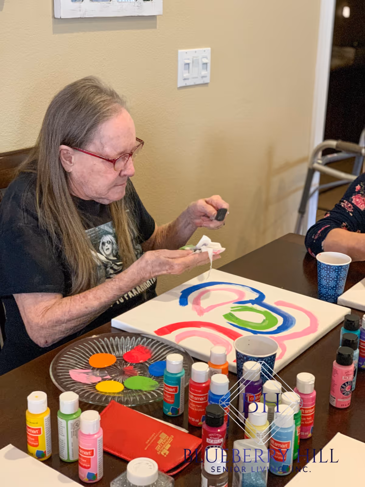 An elderly woman with long gray hair and red glasses is sitting at a table painting on a canvas. Various bottles of colorful paint and a palette with paint blobs are on the table. Another person is partially visible sitting next to her. The setting appears to be indoors with beige walls.