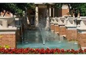 A decorative outdoor water fountain with a central jet spraying water upwards, surrounded by brick and stone architectural features, greenery, and red flowers in the foreground.