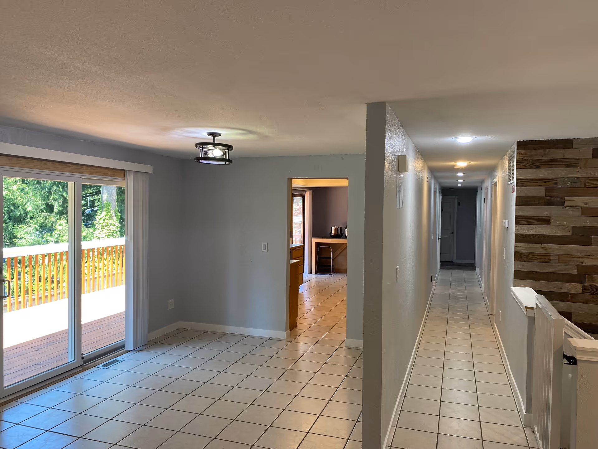 Interior view of a residential space with a tiled floor, a sliding glass door leading to a wooden deck outside, a ceiling light fixture, and a hallway with multiple doors and recessed lighting. The walls are painted light gray, and there is a wooden accent wall on the right side near the hallway.