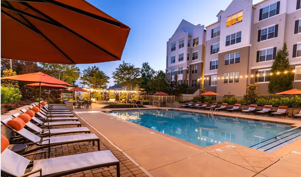 Outdoor swimming pool area at dusk with lounge chairs lined up on both sides, each with orange cushions and umbrellas. String lights are hung above a seating area in the background, and a multi-story building is visible behind the pool.