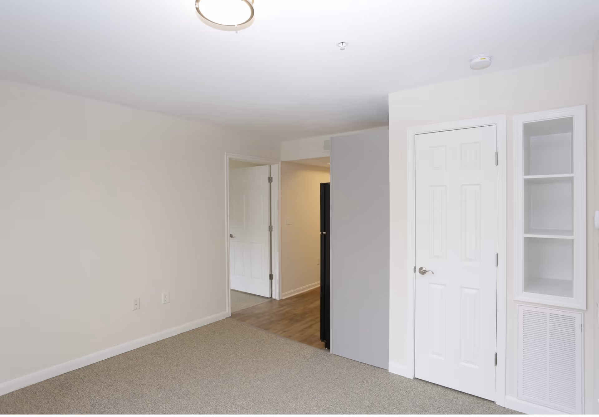 Empty interior room with beige carpet, white walls, a closed door and built-in shelving, and an open doorway to a hallway.