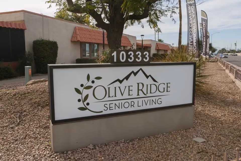 Outdoor view of Olive Ridge Senior Living facility sign with the address number 10333 displayed above it. The sign features a mountain logo and olive branch design. Behind the sign, there are single-story buildings with red-tiled awnings, trees, and a sidewalk along a street.