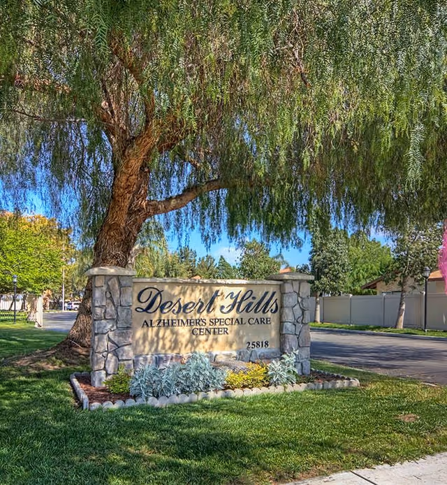 Stone sign for Desert Hills Alzheimer's Special Care Center with landscaping and a large tree behind it, situated near a road with a fence and trees in the background under a clear blue sky.