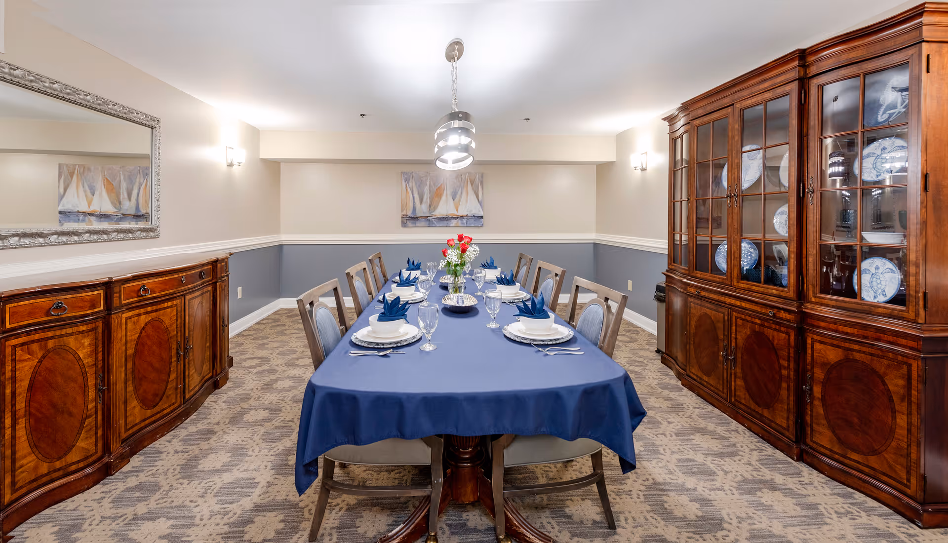 A formal dining room with a long table covered with a blue tablecloth set for eight people with white plates, blue napkins, and glassware. The room features wooden furniture including a sideboard on the left and a china cabinet on the right displaying blue and white dishes. There is a large mirror on the left wall and a painting of sailboats on the back wall. The room is softly lit with wall sconces and a modern pendant light above the table.