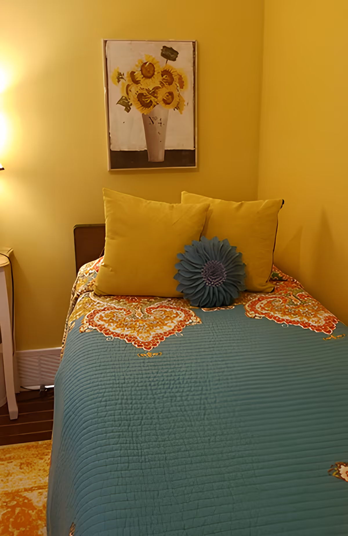 Single bed in a yellow-painted room with a teal quilt, yellow pillows and a round decorative cushion, topped by a sunflower painting.