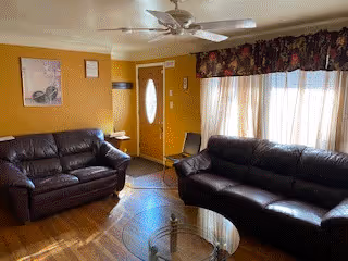 A cozy living room with two dark brown leather sofas facing each other, a glass coffee table in the center, a ceiling fan with lights, and large windows with floral curtains allowing natural light to fill the room. The walls are painted a warm yellow color, and there is a white front door with an oval window.