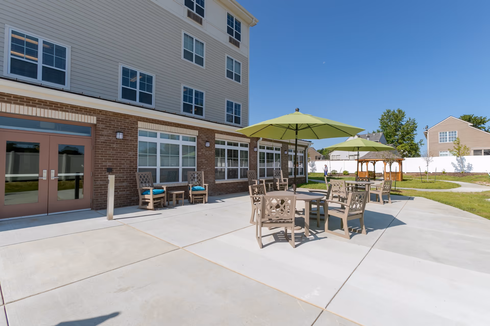 Outdoor patio area of a senior living facility with several tables and chairs under green umbrellas. The building has large windows and a brick lower facade with beige siding above. There is a gazebo and some greenery in the background under a clear blue sky.