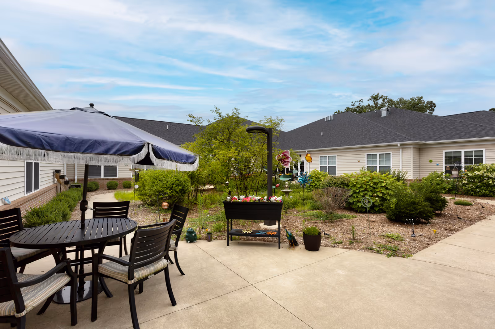 Outdoor patio area at StoryPoint Portage with a round table and four chairs under a large blue umbrella. Surrounding the patio are garden beds with various plants and shrubs, and beige buildings with dark roofs in the background under a partly cloudy sky.