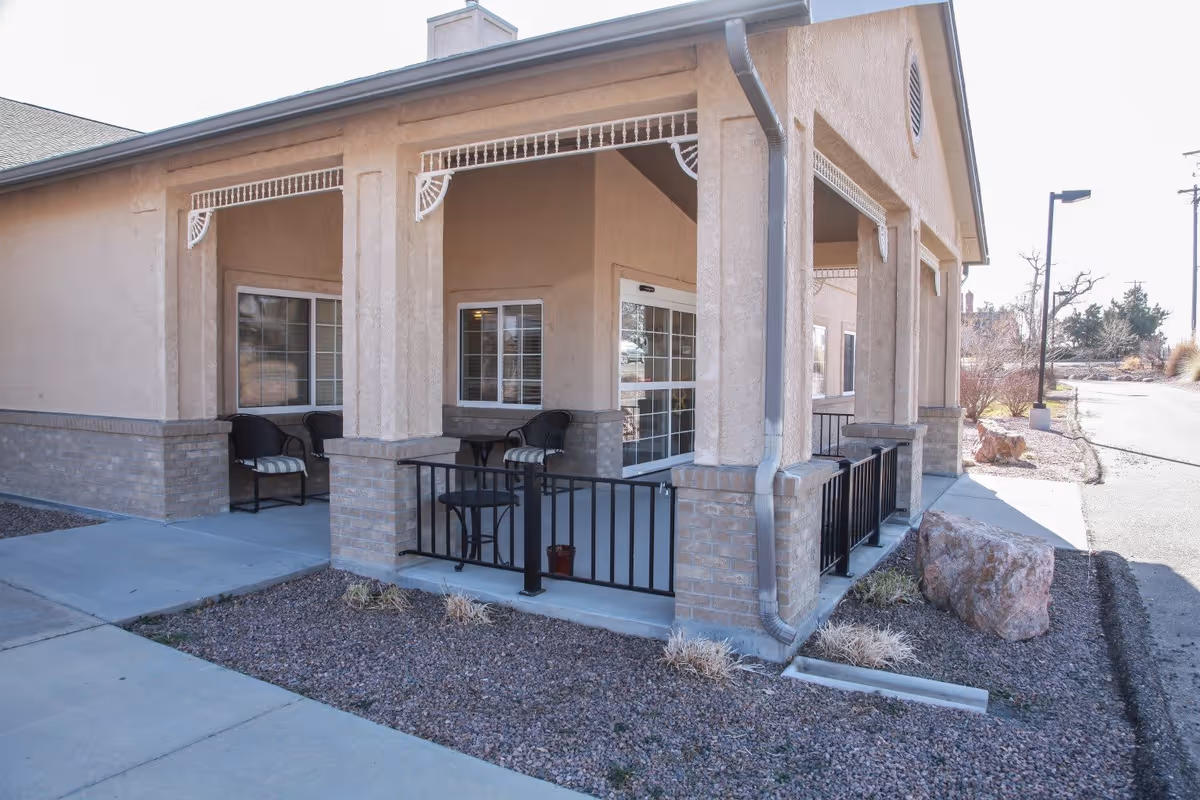 Exterior view of a covered patio area at Cottonwood Ridge facility with beige stucco walls, brick pillars, black metal railing, and outdoor seating including chairs and a small table. The patio is adjacent to a sidewalk and landscaped with rocks and sparse vegetation.