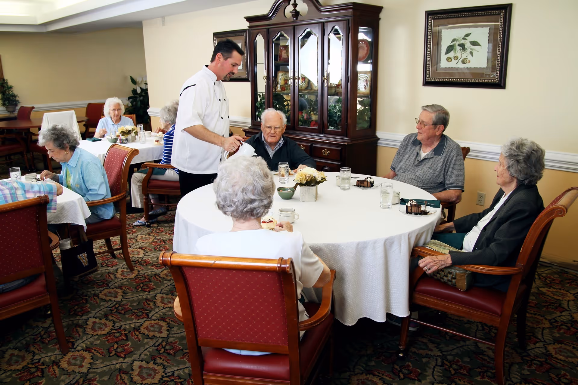 A group of elderly people sitting around a round table in a dining room, being served by a staff member in a white uniform. The room has a patterned carpet, wooden chairs with red cushions, a china cabinet, and framed artwork on the wall.