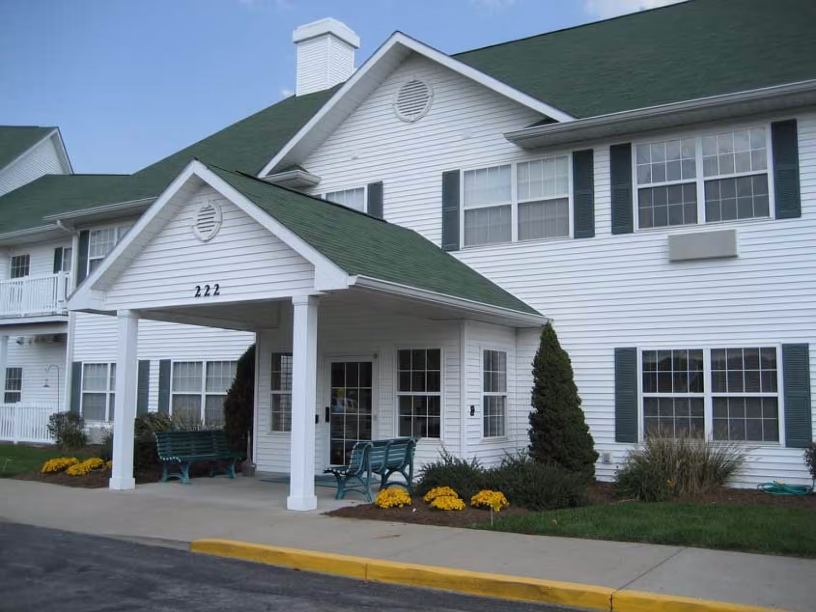 Exterior view of a white two-story building with green roof and shutters, featuring a covered entrance with the number 222 above it, green benches, and landscaping with yellow flowers and shrubs.