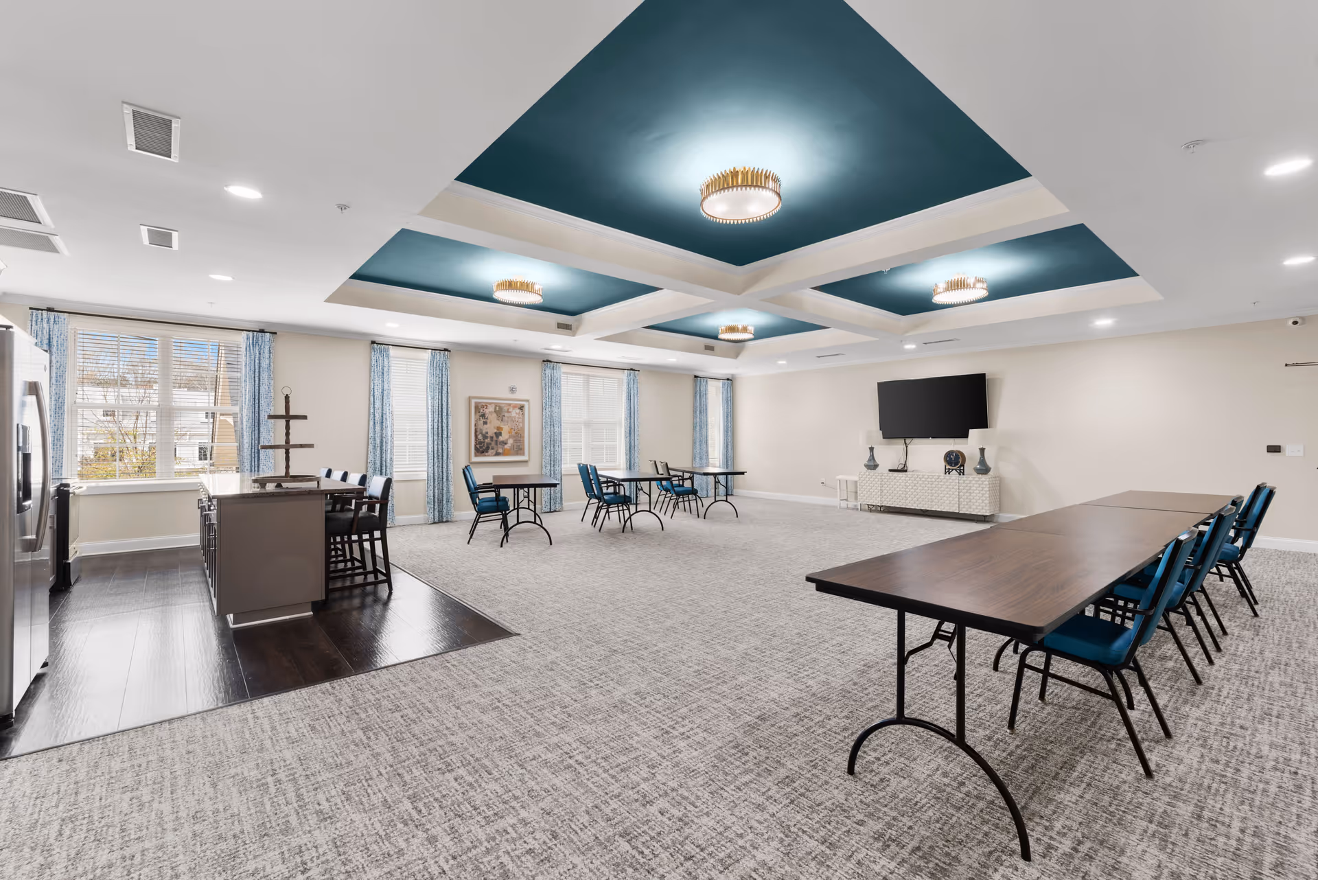A spacious, well-lit community room with a kitchen area on the left featuring stainless steel appliances and an island with bar stools. The room has multiple tables with blue chairs arranged throughout the carpeted area. The ceiling has a coffered design with teal inset panels and modern light fixtures. Large windows with blue patterned curtains allow natural light to fill the room. A flat-screen TV is mounted on the far wall above a white console table with decorative lamps and a clock.