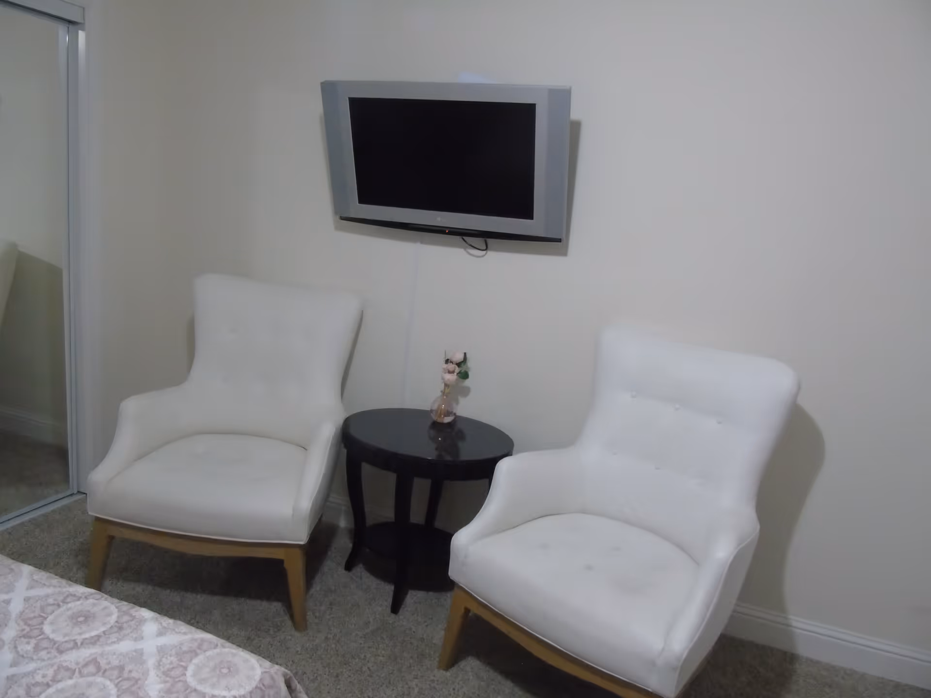 Two white cushioned armchairs with wooden legs positioned on either side of a small round black table with a small flower vase on top, a flat-screen TV mounted on a beige wall above the table, and a mirrored closet door to the left.