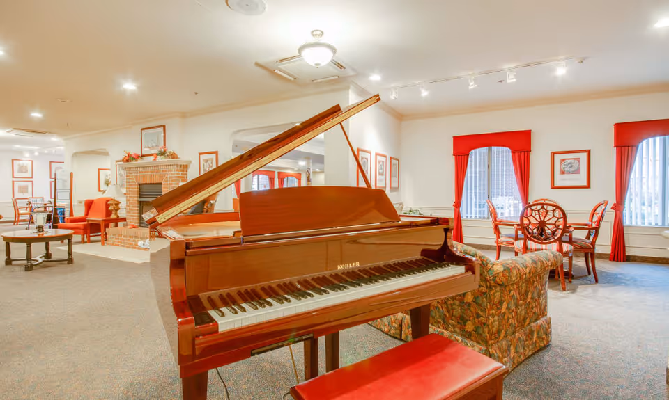 A spacious living room area in a senior living facility featuring a polished grand piano with a red bench in the foreground. The room has carpeted floors, floral upholstered sofas, red curtains on windows, framed artwork on the walls, a brick fireplace, and several wooden tables and chairs arranged throughout the space.