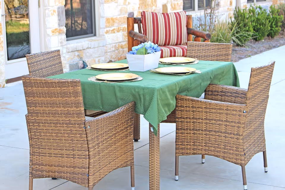 Outdoor patio dining table with a green tablecloth, wicker chairs, place settings and a small centerpiece in front of a stone building.