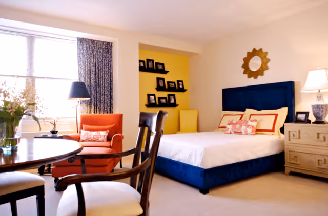 A bright and cozy bedroom featuring a blue upholstered bed with white and red pillows, a wooden nightstand with a lamp, a round wooden table with chairs, an orange armchair near a window with curtains, and a yellow accent wall with shelves displaying framed photos.