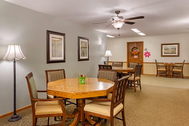 Interior view of a senior living facility common area with multiple wooden tables and chairs arranged for seating. The room is softly lit with floor and table lamps, decorated with framed artwork on the walls, and has a ceiling fan with a light fixture. The carpeted floor and neutral wall colors create a warm and inviting atmosphere.