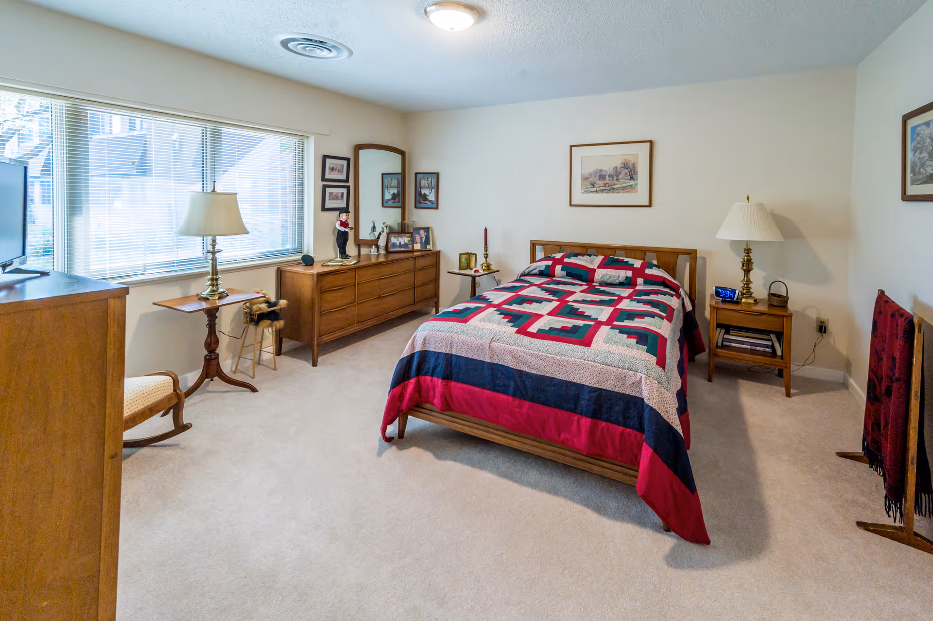 A bright bedroom with a bed covered by a red, navy, and green patchwork quilt, wooden dressers and nightstands with lamps, and a large window.