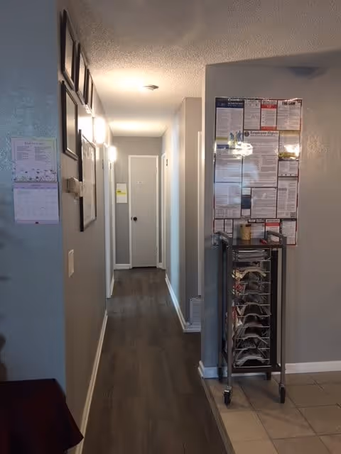 A narrow hallway in a residential or care facility with light gray walls and dark wood flooring. On the right side, there is a bulletin board with various notices and a metal rack holding papers or magazines. The hallway leads to a closed white door at the end, with additional doors on the right side. The ceiling has a light fixture, and there are framed pictures on the left wall.