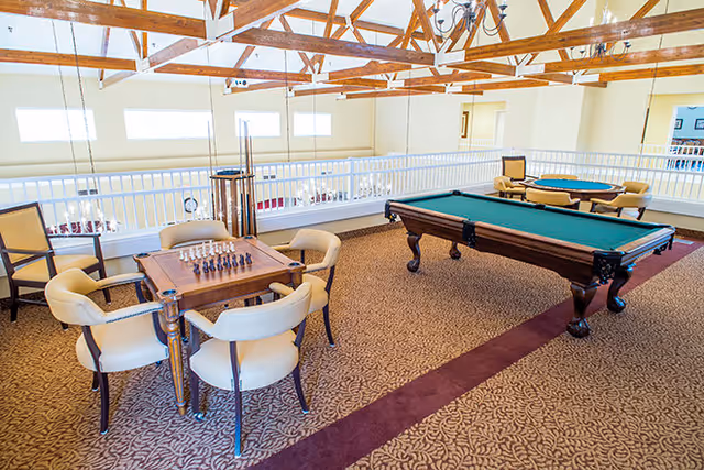Bright open recreation room with a pool table, a chess table surrounded by chairs, and exposed wooden ceiling beams.