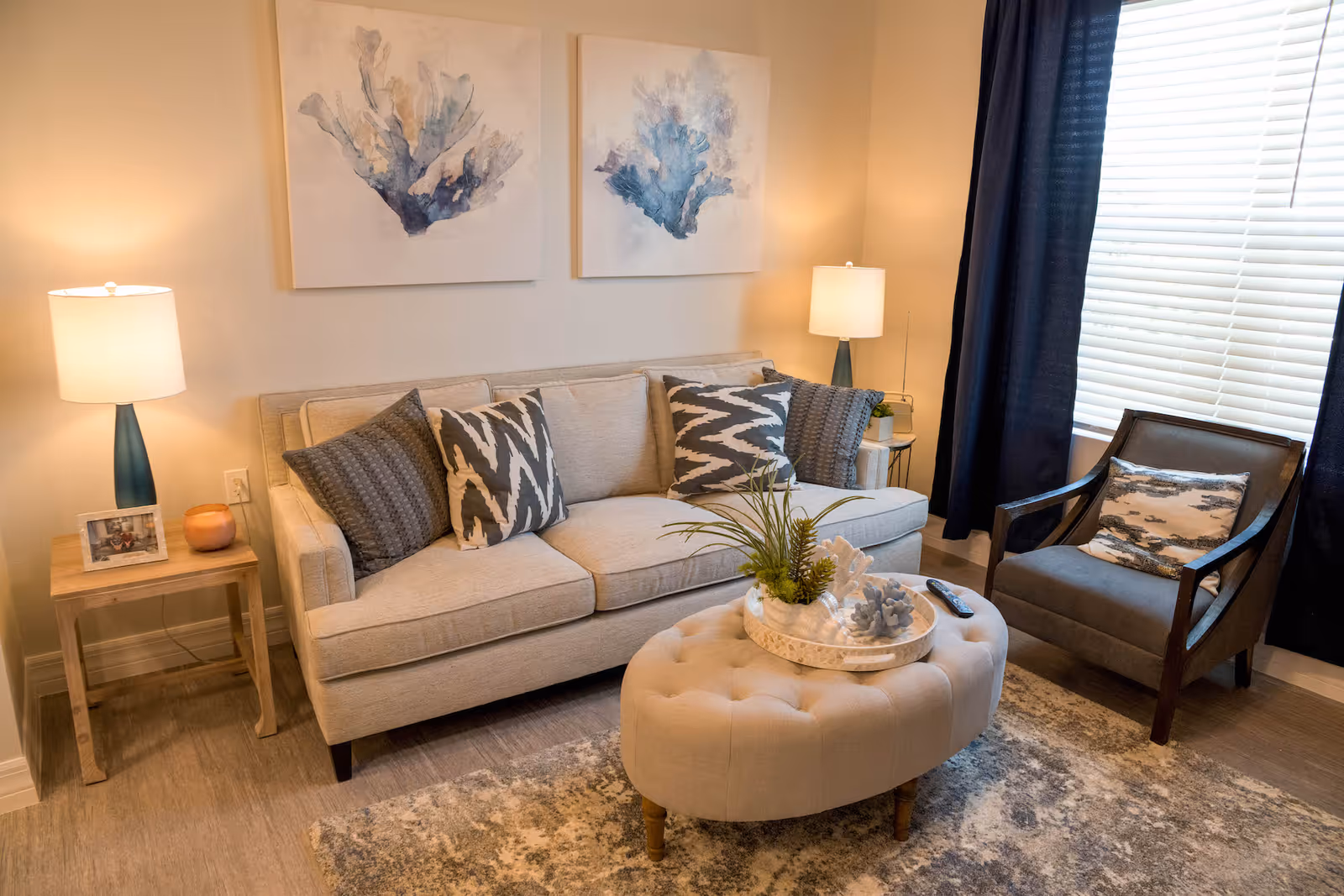 A cozy living room with a beige sofa, patterned throw pillows, an upholstered round ottoman, an armchair, side tables with lamps, and abstract wall art by a window.