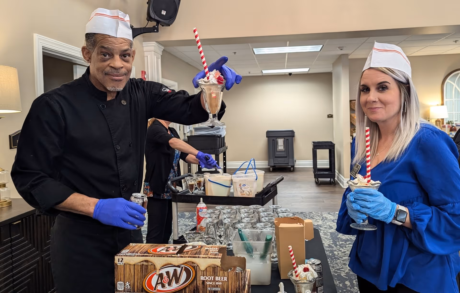Two staff members wearing white hats and blue gloves hold root beer floats with whipped cream and cherries in a senior living facility dining area. One man in a black chef coat holds up a float and a can of A&W root beer, while a woman in a blue dress smiles holding her float. Behind them, another person prepares more floats on a table with glasses and ingredients.