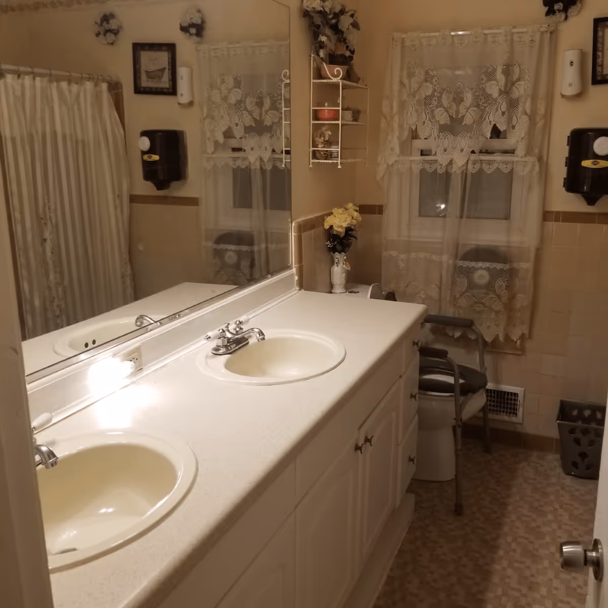 A bathroom with a double sink vanity, a large mirror above the sinks, a window with lace curtains, a shower with a striped curtain, a toilet with a raised seat and armrests, a wall-mounted soap dispenser, and decorative flowers on the wall and countertop.