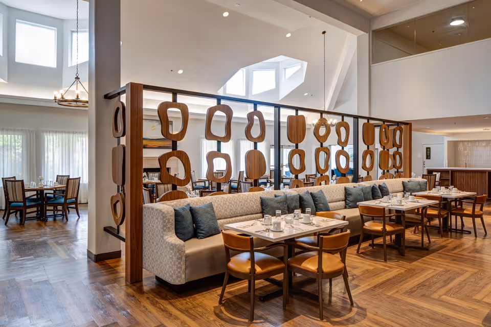 A spacious and well-lit dining area with wooden floors, tables set with glasses and napkins, cushioned bench seating with blue pillows, and wooden chairs. Decorative wooden panels separate different sections of the room. Large windows with sheer curtains allow natural light to fill the space.