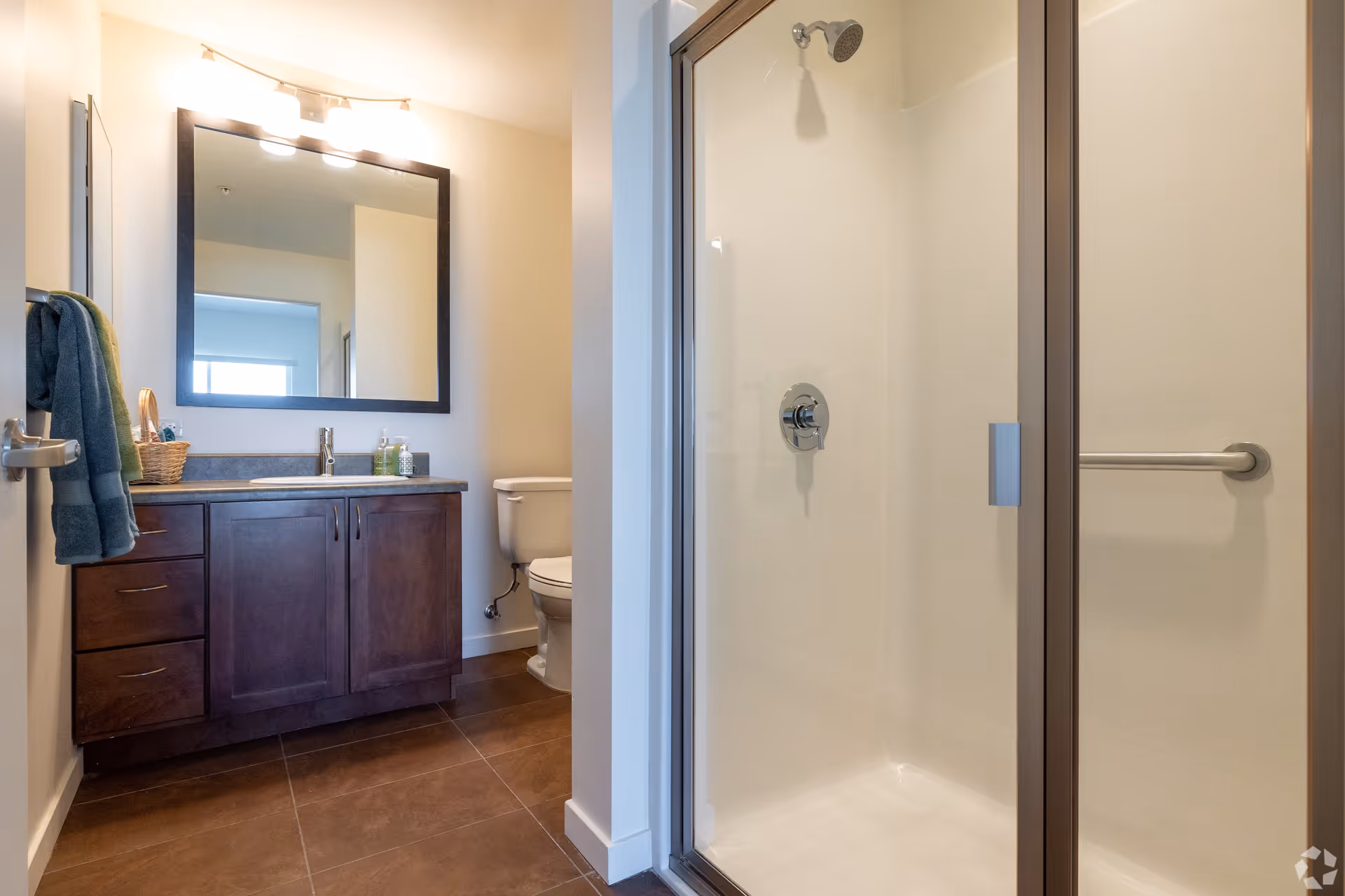 A clean and modern bathroom featuring a glass-enclosed shower with a metal grab bar, a toilet, and a vanity with a dark wood cabinet, a sink, and a large mirror above it. Towels hang on a rack near the door.