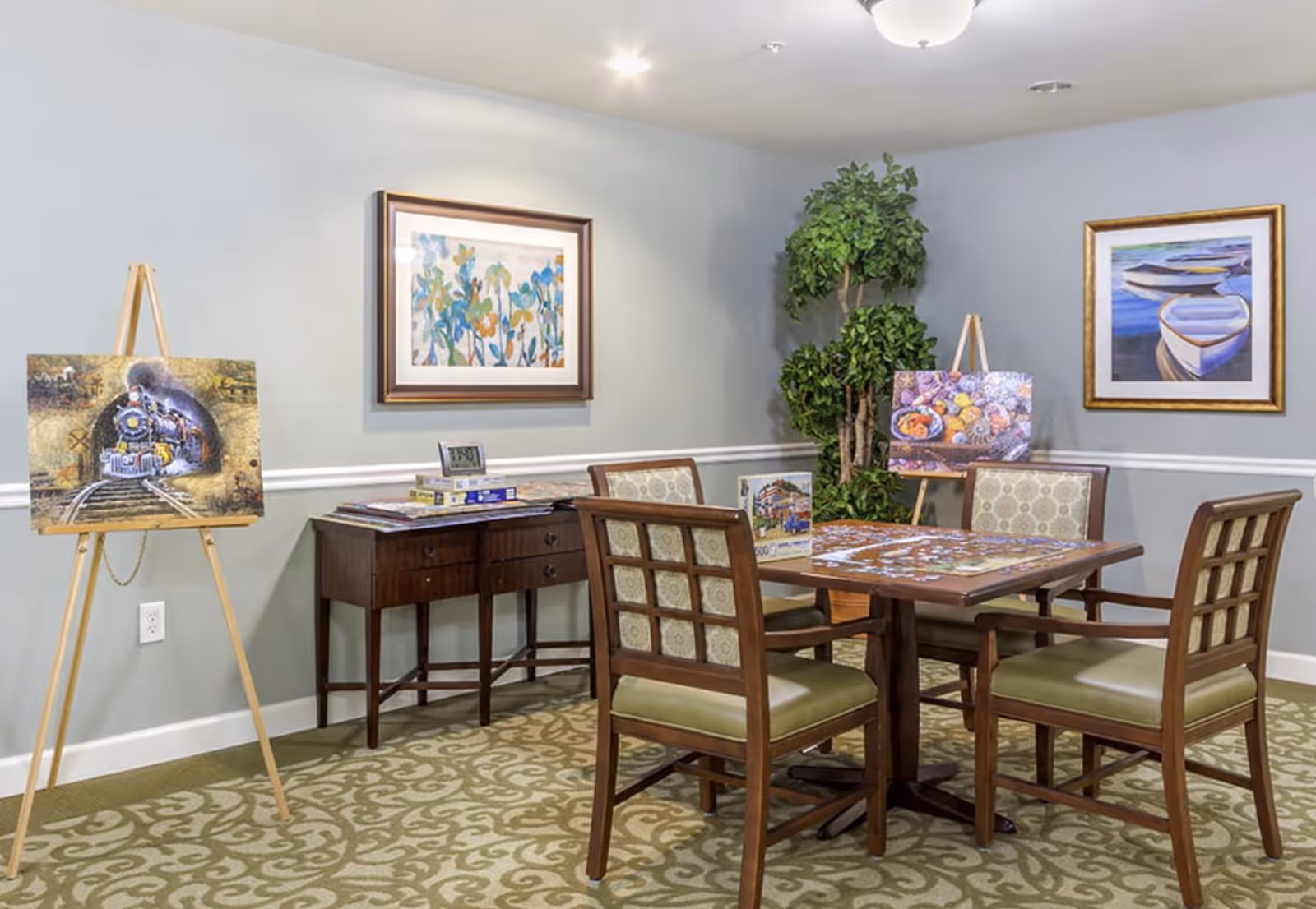 A cozy interior room with a square wooden table surrounded by four cushioned chairs. The table has a partially completed jigsaw puzzle on it. The room has light gray walls adorned with framed artwork and two easels displaying paintings. A small wooden side table holds board games and a digital clock. A tall green plant is placed in the corner.