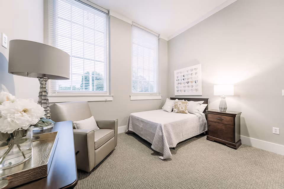 A bright and clean bedroom in a senior living facility featuring a bed with white and beige bedding, a wooden nightstand with a lamp, a comfortable armchair, two large windows with white blinds, and a decorative framed artwork on the wall.