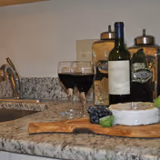 Close-up view of a kitchen countertop with a bottle of red wine, two filled wine glasses, a wooden cutting board with cheese, grapes, and a cucumber slice, and two glass jars filled with dry food items in the background near a sink.