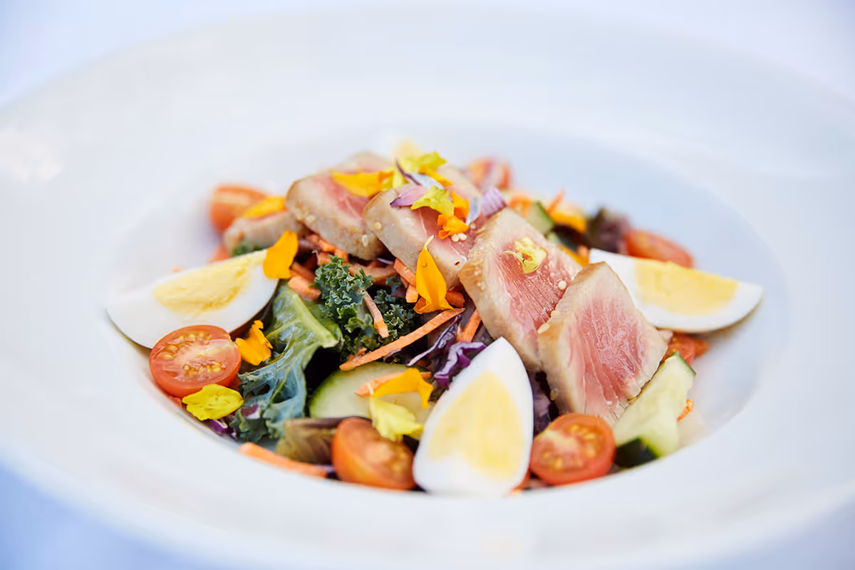 A close-up of a fresh salad served in a white bowl, featuring slices of seared tuna, quartered hard-boiled eggs, cherry tomato halves, cucumber slices, leafy greens, shredded carrots, and edible flower petals.