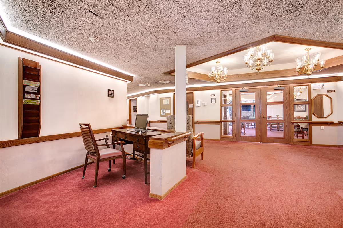 Interior view of a senior living facility lobby or reception area with a wooden desk and chair, a computer on the desk, red carpet flooring, white walls with wooden trim, chandeliers hanging from the ceiling, and glass double doors leading to another room with tables and chairs.